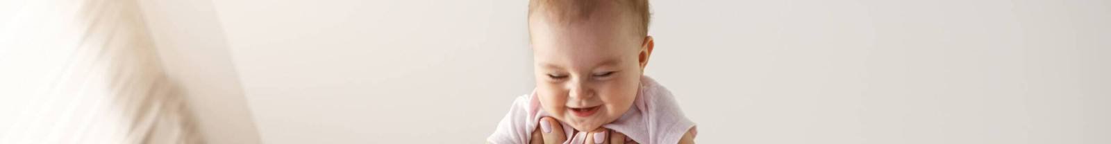 Young cheerful mother playing with her newborn baby lying on bed at home. Copy space.